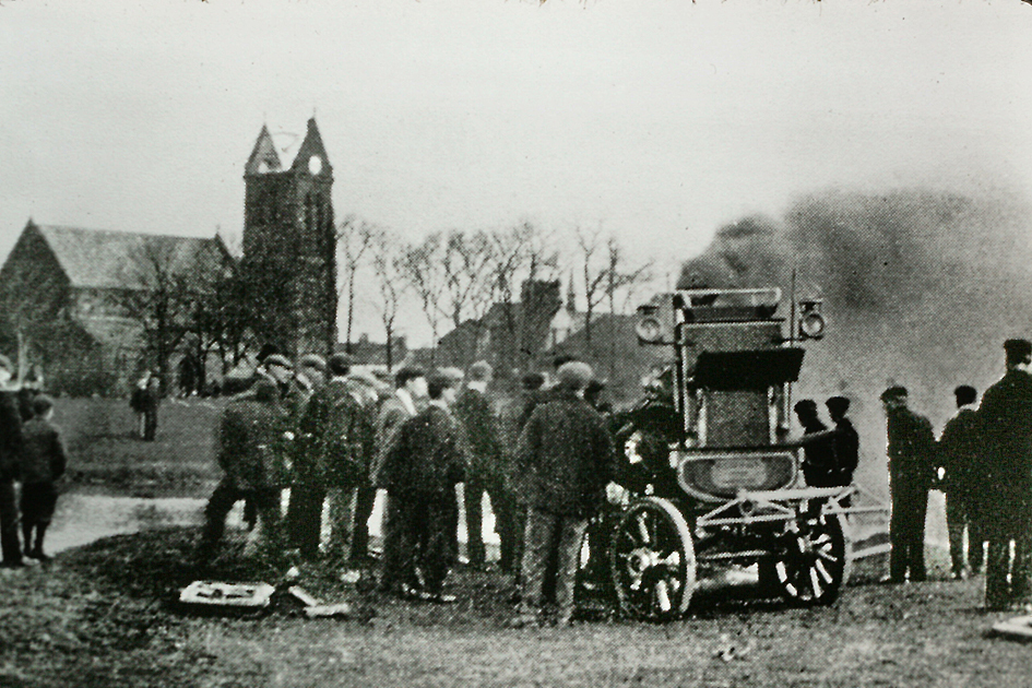 Horse drawn fire pump with volunteers and spectators by the pond on Half Acre Green in Marske with the burnt out church tower in the background