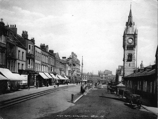 Darlington and Stockton Times: High Row on a sunny day that has caused all of the shops to pull down their blinds so their window goods were not bleached or damaged. On the right, the cabs are parked outside the market hall, and there is shelter in the street for the cabmen